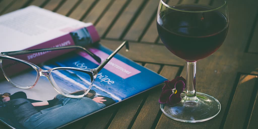 Glas Rotwein neben Buch und Lesebrille auf einem Holztisch